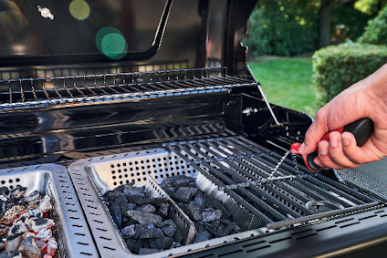 Hand håller termometer över en gasolgrill med kolbriketter och träflis i metallfack.