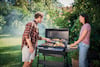 Man och kvinna grillar mat på en kolgrill i en grönskande trädgård. Rök stiger från grillen.