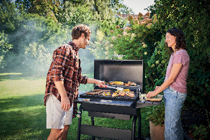Man och kvinna grillar mat på en kolgrill i en grönskande trädgård. Rök stiger från grillen.