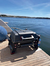 En bärbar grill står på ett träbord, med utsikt över ett lugnt vatten och klippiga stränder under en klar blå himmel.