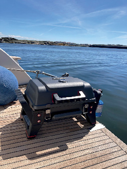 En bärbar grill står på ett träbord, med utsikt över ett lugnt vatten och klippiga stränder under en klar blå himmel.