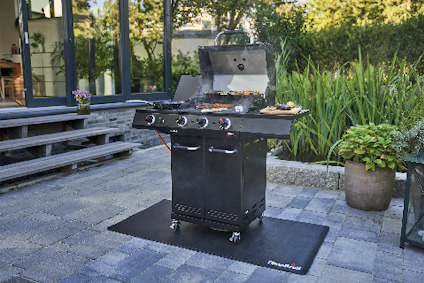 Utomhusgrill på stenlagd uteplats, omgiven av grönska och trämöbler, med diverse grillredskap på sidohyllan.