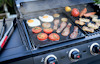 Gassgrill med grillbricka, steker ägg, tomater, bacon, paprika och korv. Reglageknappar synliga på framsidan.