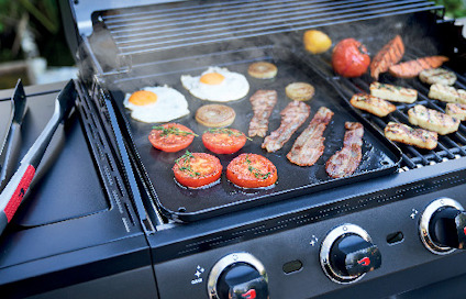Gassgrill med grillbricka, steker ägg, tomater, bacon, paprika och korv. Reglageknappar synliga på framsidan.