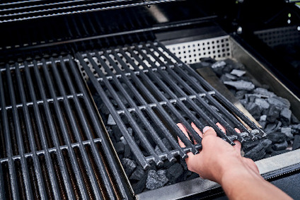 En hand lyfter ett grillgaller över grillkol i en utomhusgrill.