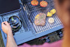 En person lagar mat på en grill med kött, majskolv, tomat och bröd. En liten kastrull används i sidan av grillen.