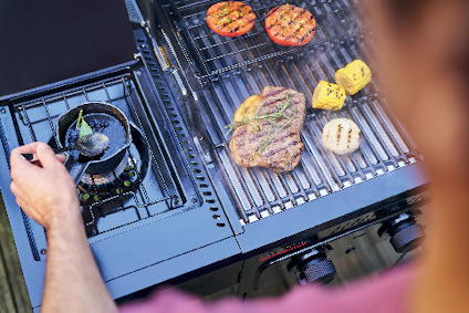 En person lagar mat på en grill med kött, majskolv, tomat och bröd. En liten kastrull används i sidan av grillen.