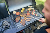 Grillad mat inklusive kött, korvar och grönsaker på en utomhusgrill. En person hanterar maten med en tång.