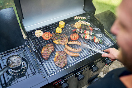 Grillad mat inklusive kött, korvar och grönsaker på en utomhusgrill. En person hanterar maten med en tång.