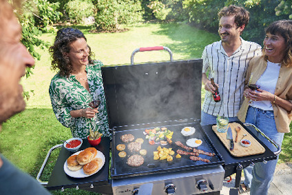 Fyra personer runt en grill med hamburgare, korv, grönsaker, bröd och dryck. Två av dem håller i glas.