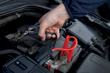 Hand håller i en tång över ett bilbatteri med en röd kabelklämma och flera anslutningar.