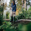 Person i stövlar räfsar i en grönskande trädgård med växthus i bakgrunden.