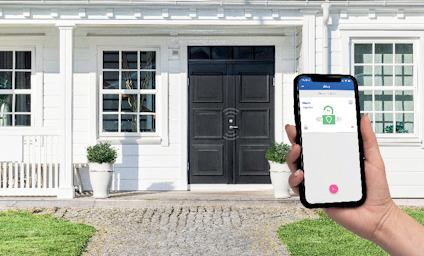 Hand håller en mobiltelefon med en app, framför ett vitt hus med svarta dörrar och en stenlagd gång.