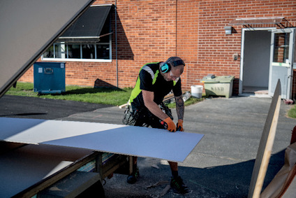 Person i skyddsutrustning arbetar med byggmaterial utomhus framför en tegelbyggnad.