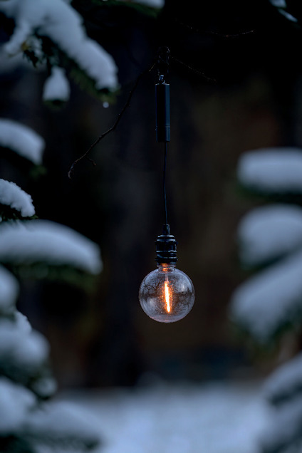Glödlampa hänger utomhus mellan snötäckta grenar.