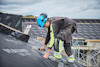 Person i blå hjälm installerar takmaterial på sluttande tak med utsikt över ställningar och molnig himmel.