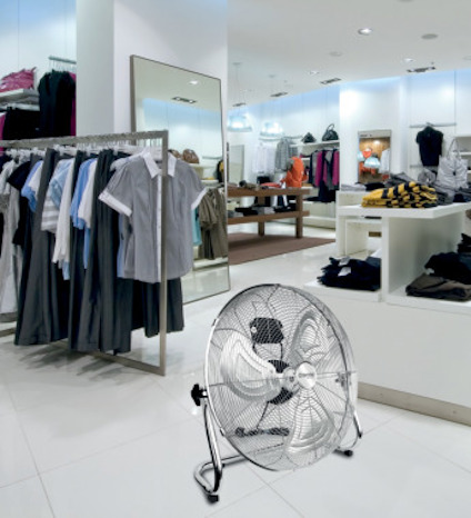 Fläkt med stort galler står på vit golvyta i en klädbutik med speglar och hängkläder.