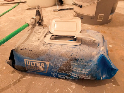 Blå förpackning med våtservetter, texten "Ultra Grime", liggande på ett golv med verktyg och målarburk i bakgrunden.