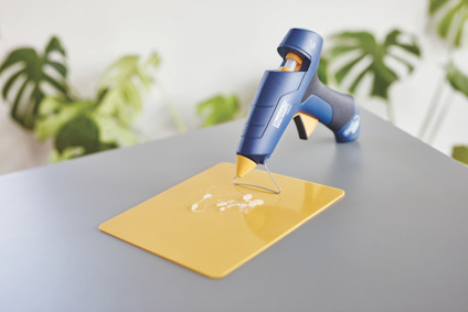 Limpistol på ett bord bredvid ett gult underlägg med lim. Bakgrund med gröna växter.
