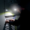 Person justerar brytare i ett elektriskt skåp, bär pannlampa i mörk miljö.
