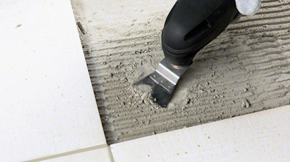 En person använder en platt spackelspade för att applicera eller jämna ut grått murbruk på kakelunderlag.